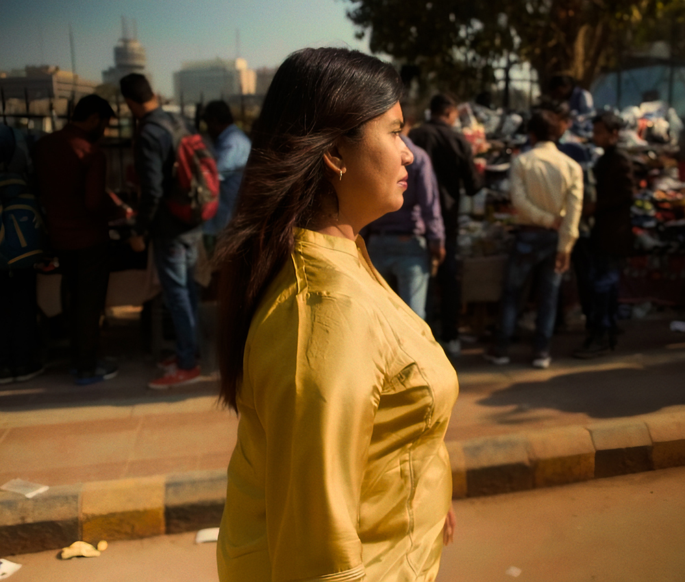 A woman in profile wearing a yellow shirt walks down a city street.